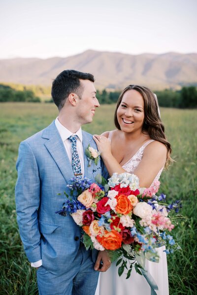 A man and woman in wedding attire smiling while standing in a field in the Cades Cove loop holding a big bouquet of colorful flowers.
