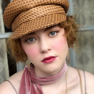 A girl with red curly hair wearing a straw hat and pink scarf  and blushed lips looks pensive