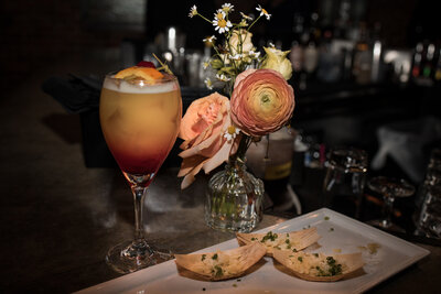 vignette of signature drink with a flower vase with peach flowers next to a trio of appetizers served in canoe shaped wooden boats.
