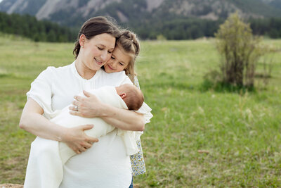 Mom holding and looking at her newborn baby. Behind her her daughter is hugging her and looking at the baby