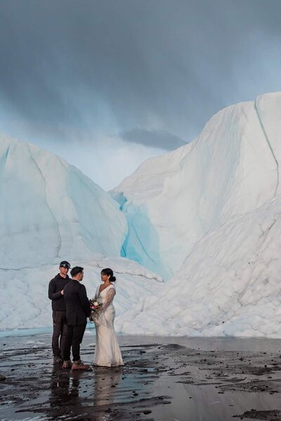 A bride and groom stand in front of a huge wall of white ice during their private helicopter elopement on Matanuska glacier in Alaska.  The officiant is standing nex to them and is holding a vow book, but they are far away so look tiny against the wall of glacier ice and snow.