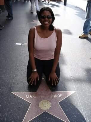 Catherine kneels and smiles beside Marilyn Monroe's star on the Hollywood Walk of Fame, wearing a pink tank top and black pants.
