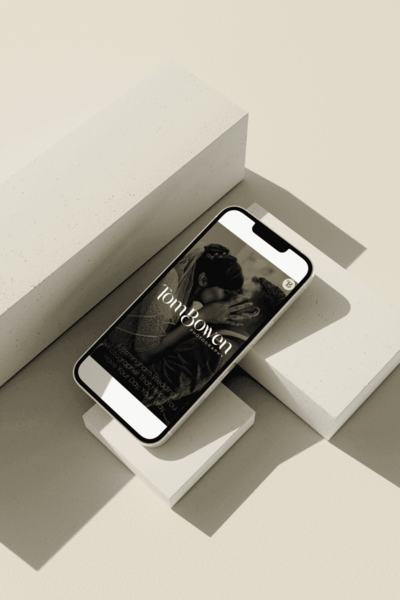 A smartphone displaying the homepage of Tom Bowen Photography, featuring a black and white image of a bride and groom kissing. The text "Tom Bowen Photography" is prominently displayed on the screen, along with a tagline about Birmingham wedding photography. The phone is placed on a beige geometric surface with shadows, creating a modern and minimalist look.