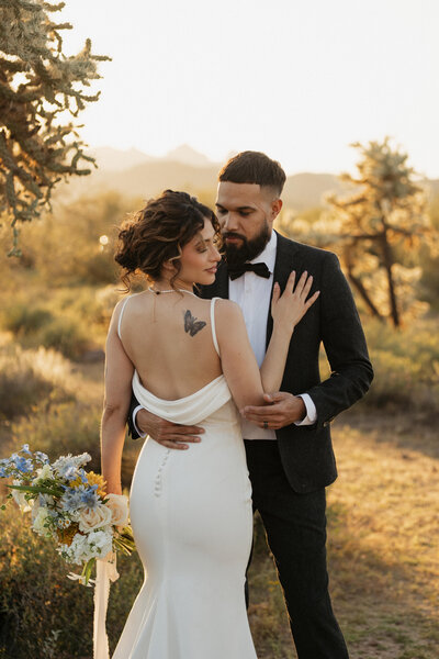 bride holding her bridal bouquet while touching groom's chest as they both stand in a field with trees in the background