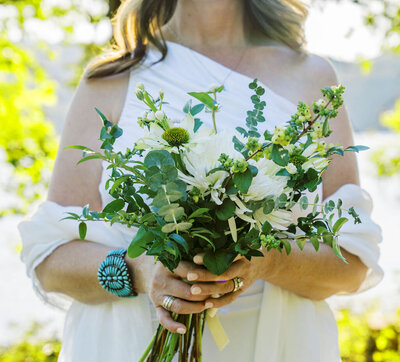 Bride holding a natural, hand-tied bouquet of white flowers and greenery, adorned with turquoise jewelry, captured by Joseph Mayden during a sunlit outdoor elopement.