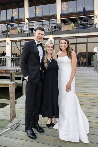 Three people stand on a wooden pier in front of a building with large windows. The man on the left wears a tuxedo, and the two women are in formal dresses. They are all smiling and posing for the photo.