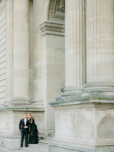 a woman and man in a black suit and black tulle dress  on some steps at the louvre looking at one another and both looking off into the distance