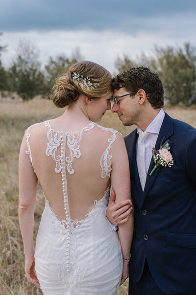 Bride and groom standing close with foreheads touching in an emotional moment; the groom gently caresses the bride’s arm as she wears a wedding dress and he wears a suit, surrounded by a grass-filled patch, captured by a Bendigo wedding and portrait photographer.