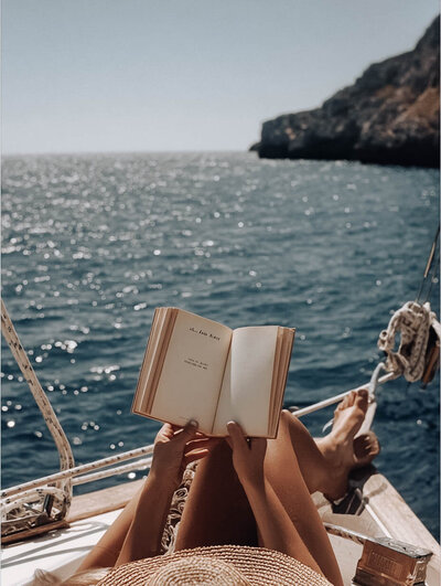 Relaxed traveler reading on a boat, soaking up the sun on the European coastline. Tailored Oasis Travel Co.