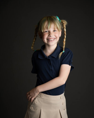Girl in a blue and khaki school uniform with blue and blonde braids dancing in her school portrait on a gray backdrop, photo by Jessica Bowles Photography & Video
