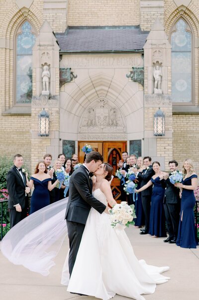 bride and groom kissing as wedding party watches