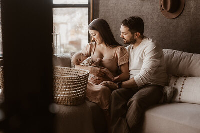 Mom and husband cuddle newborn baby girl in a dark front room with a wicker Moses basket