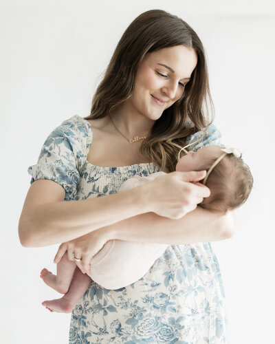 Smiling mother in a blue floral dress holding her newborn in a natural light studio, captured by a Kansas City newborn photographer.