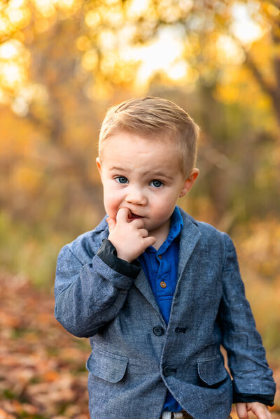 Toddler boy in blue sport jacket bites on finger and looks directly into camera with yellow fall leaves behind him.