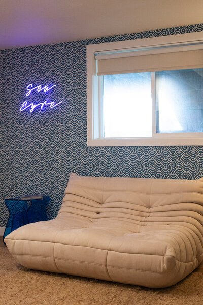 A cozy room features a modern beige futon-style couch against a blue patterned wall. Above the couch, a neon sign glows with cursive writing, and a small blue side table sits to the left.