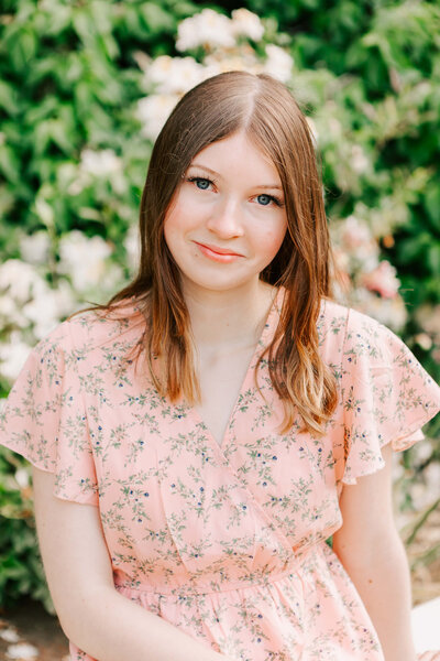 A high school girl in a pink dress sits on a bench in front of a flower bush in Salem, OR