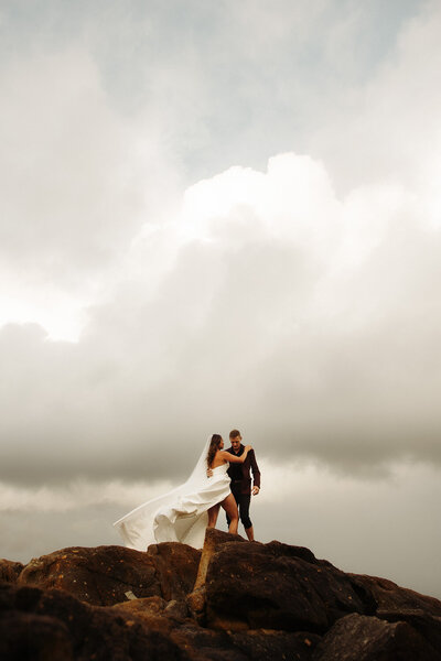 Bride and groom standing on rocky cliffs, the bride's veil blowing in the wind under a dramatic cloudy sky during an adventurous elopement.