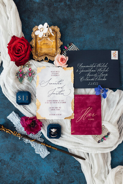 Romantic red and navy invitation suite styled on blue with florals and gold accents.