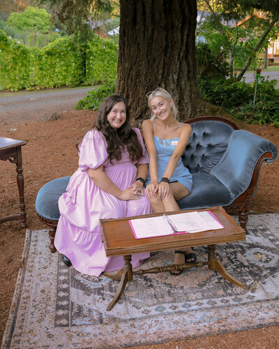 Wedding and event planners in Oregon, Clarae and Taressa, smile and sit on a lounge chair with a binder at a wedding they planned.
