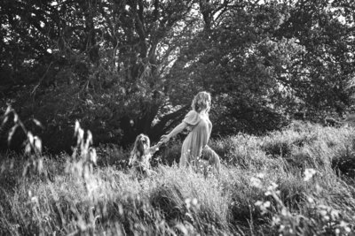 a black and white photograph of a pregnant woman leading her daughter through a field of grass with a giant tree behind them.