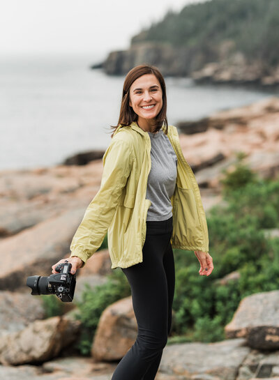 Photo of a girl holding a camera in one hand and smiling at the camera. she has a mountain behind her