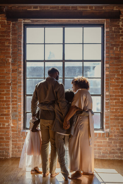 A family of four, mother, father, son, and daughter, stand closely together facing a large industrial window in a brick studio. Captured from behind, they hold each other in a quiet, connected moment bathed in natural light.