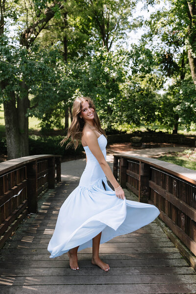 A woman in a white dress sits on a ledge, smiling and holding a graduation cap aloft, surrounded by vibrant red and yellow tulips, with trees in the background.