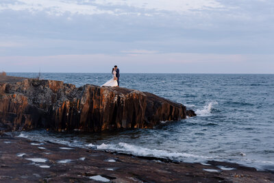 Couple holds each other on a golden-lit cliff, with waves crashing beneath them during their once-in-a-lifetime elopement.