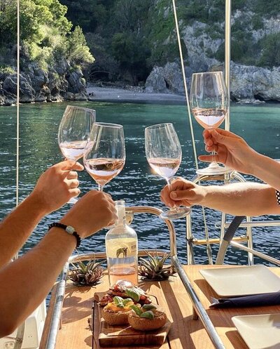 Friends toasting wine glasses over waterfront lunch during intimate group sailing experience. Tailored Oasis Travel Co.
