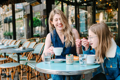 Mother and daughter at a Parisian café near the Eiffel Tower — mother laughing as her daughter takes a sip of her drink during a joyful family photoshoot.
