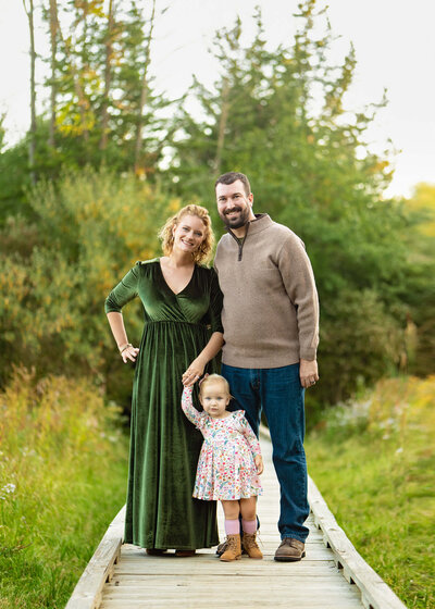 A  mom and dad stand on a wooden walkway with theiir toddler daughter, smiling