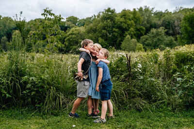 Three children stand in a lush green field, huddled together in a group hug, smiling and enjoying each other's company.
