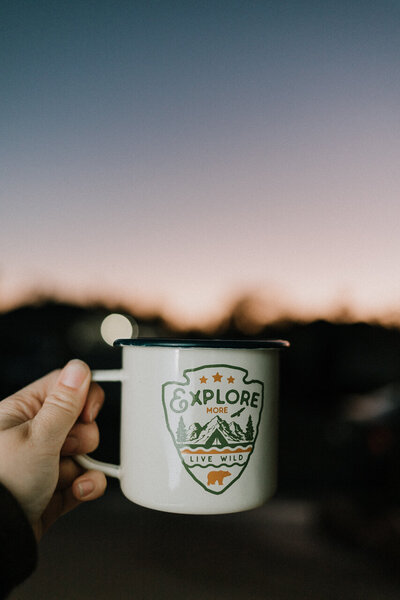 A mug that says "explore more" is held up to the sunrise