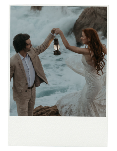 Bride and groom hold a lantern together while bride holds dress with other hand behind them is the ocean