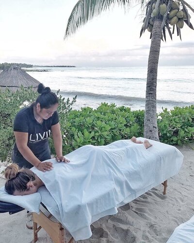 A massage therapist from Livin Massage in Tulum providing a relaxing body treatment on the beach, under palm trees with an ocean view, blending natural serenity with healing touch.