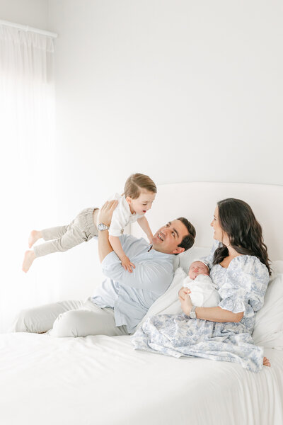 Parents sit on a bed as dad makes toddler son fly and mother holds sleeping newborn