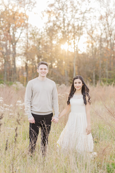 A newly engaged couple hold hands while standing in a field of tall golden grass at sunset