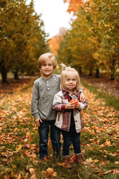 Brother and sister smiling at the camera in a Michigan orchard during fall.