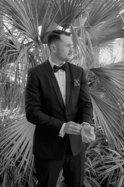 Black and white photo of groom standing in front of palm leaves adjusting his cufflinks