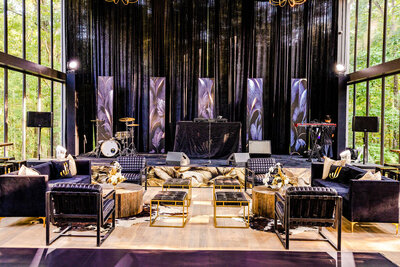 Chic black and gold event lounge with modern velvet seating, wood accents, and a dramatic stage featuring custom backdrops, drum kit, DJ booth, and keyboard setup, surrounded by floor-to-ceiling windows overlooking a forested backdrop, styled by Wells Design Co.