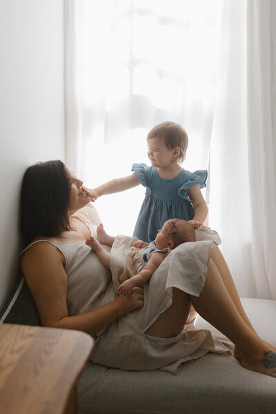 Mother sitting next to window with her toddler daughter and newborn daughter, photographed by Jacoby Andrick