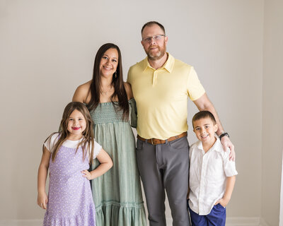 family of four standing in a studio, looking at the camera smiling.  mom dad are standing in the back with arms around each other, son has a hand in his pocket leaned against dad, and daughter has her hand on her hip standing partially in front of mom