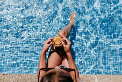 A woman in a black bathing suit and sunglasses sits at the edge of a sparkling blue pool while dipping her heet in the water and holding a yellow drink.