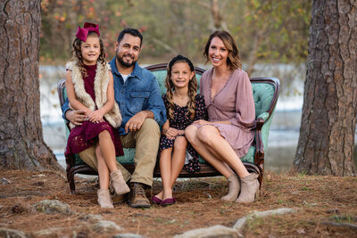 A family of four is sitting on a green chair in between two trees as they pose for their family photoshoot with Ashley Zohil Photography.