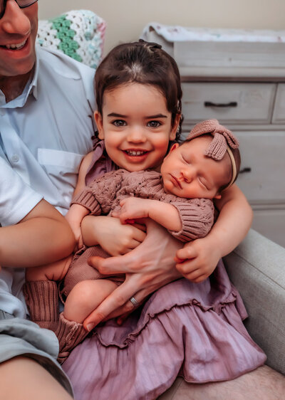 A family of a mom, dad, two toddler girls, and a dog sit together on rocks on the edge of a creek with fall colored trees behind them. They're all looking down at the tan and white dog smiling while wearing fall colors.