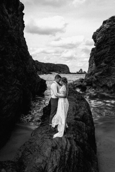 M & J facing one another holding each other tight while standing on a rock surrounded by ocean and rock formations on each side of them. black n white imag
