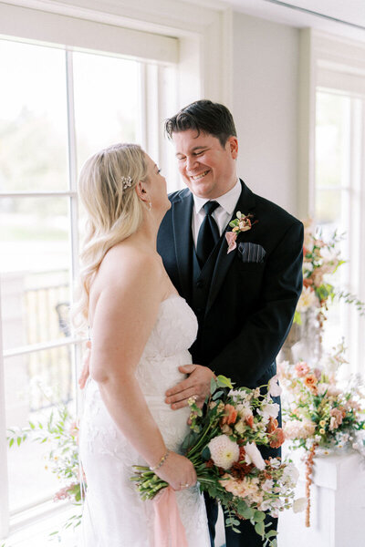 Bride and groom laughing together by the window surrounded by pastel floral decor at Oakville wedding venue
