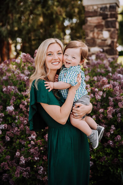 Young mom in green dress holds her baby son cheek to cheek with her in front of purple blooms.