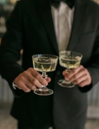 A groom in a black and white tuxedo holding two glasses of champagne.