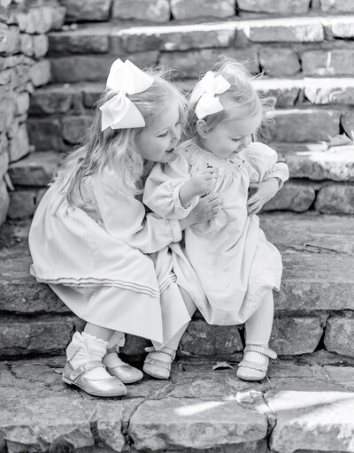 Black and white portrait of two young sisters sitting together on stone steps, dressed in classic smocked dresses with matching white bows. The older sister leans in with a smile, capturing the timeless beauty of sibling bonds in a heartfelt Birmingham family photography session